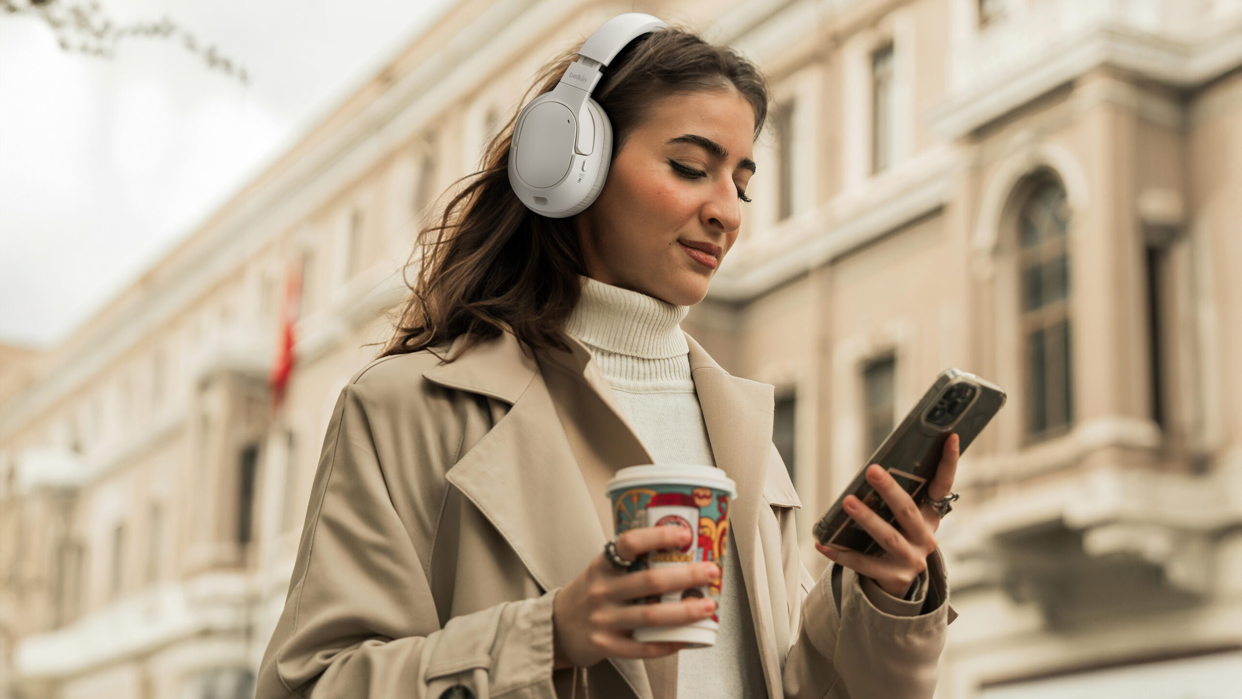 En ung kvinne i beige frakk og hvit pologenser lytter til musikk med sandfargede Belkin SoundForm Isolate hodetelefoner mens hun holder en takeaway-kaffekopp og ser på mobiltelefonen sin. Hun står utendørs foran en klassisk bygning med lyse vegger.